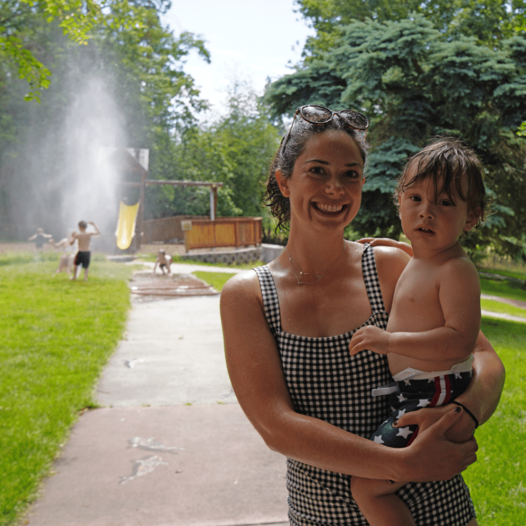 smiling woman in swimsuit holding toddler outside near sprinker